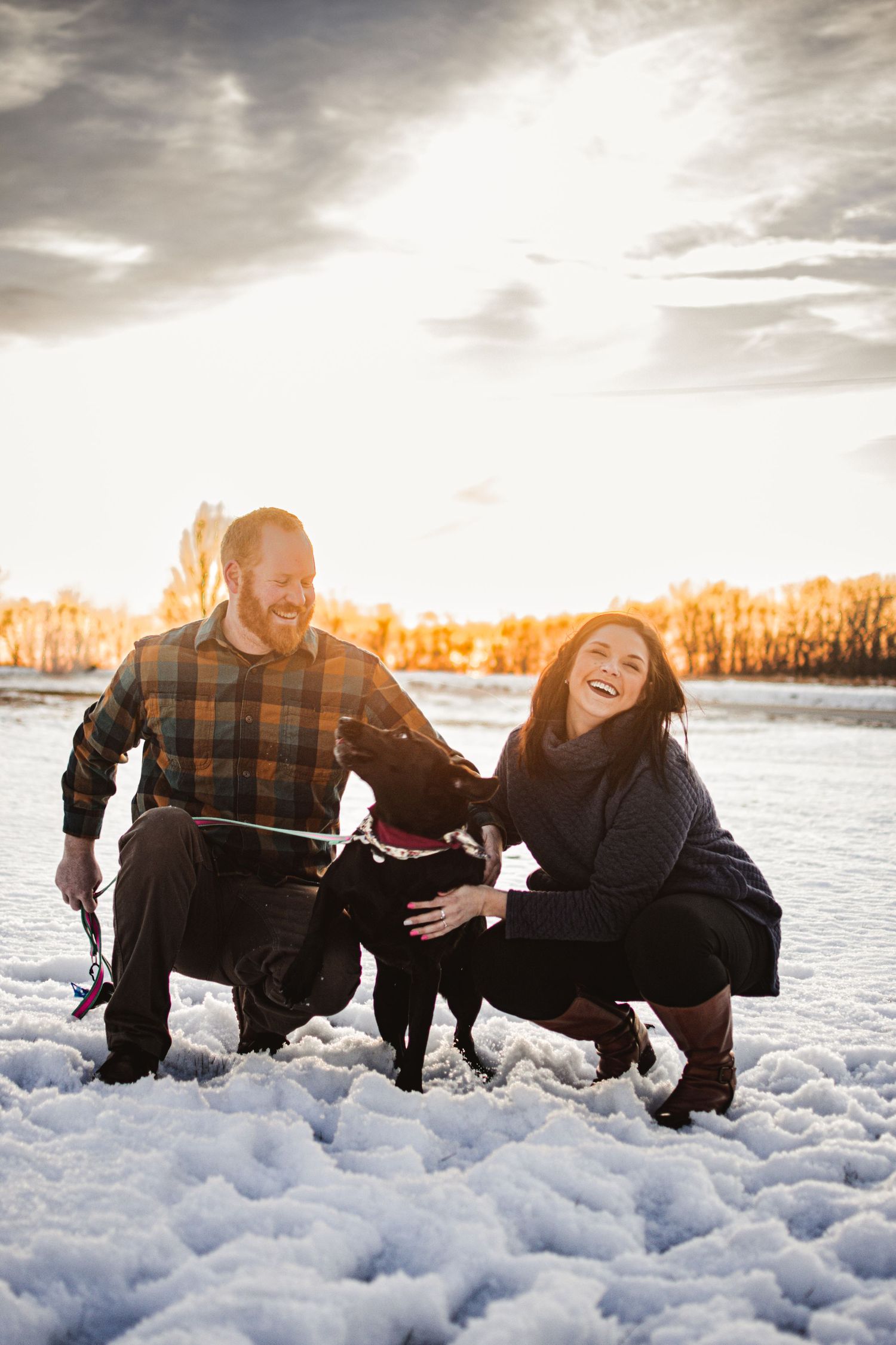 Couple and their black dog enjoying a snowy winter day at sunset.