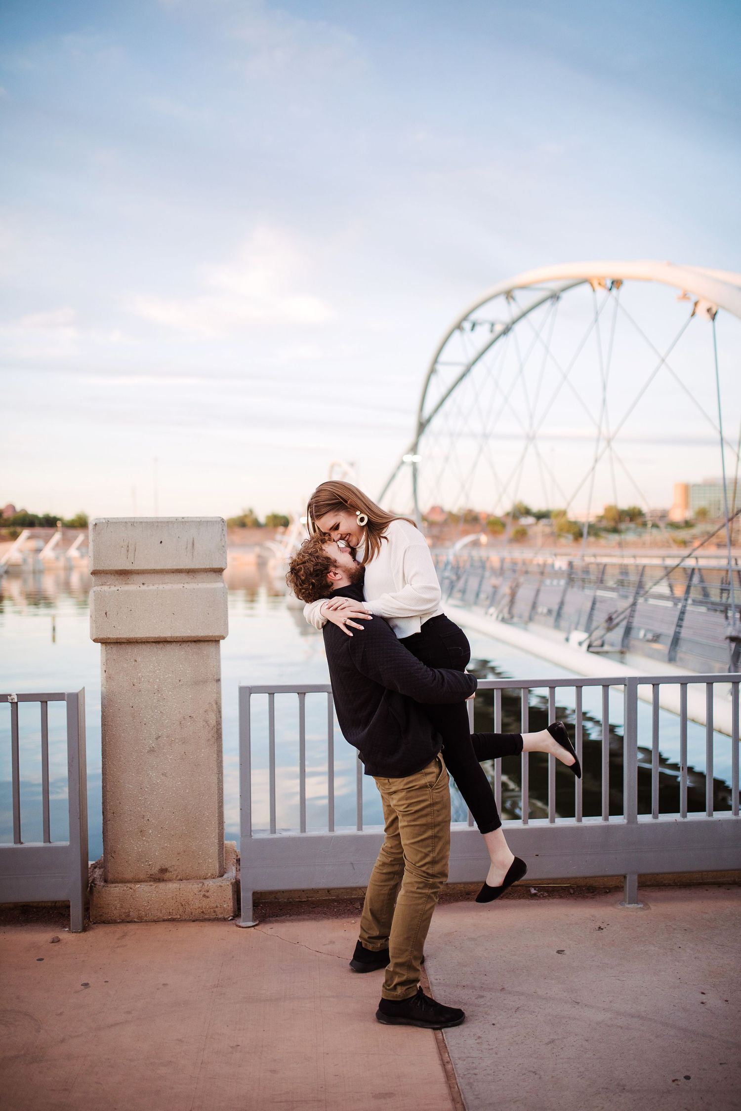 Romantic couple embracing as man lifts woman near waterfront bridge at golden hour.
