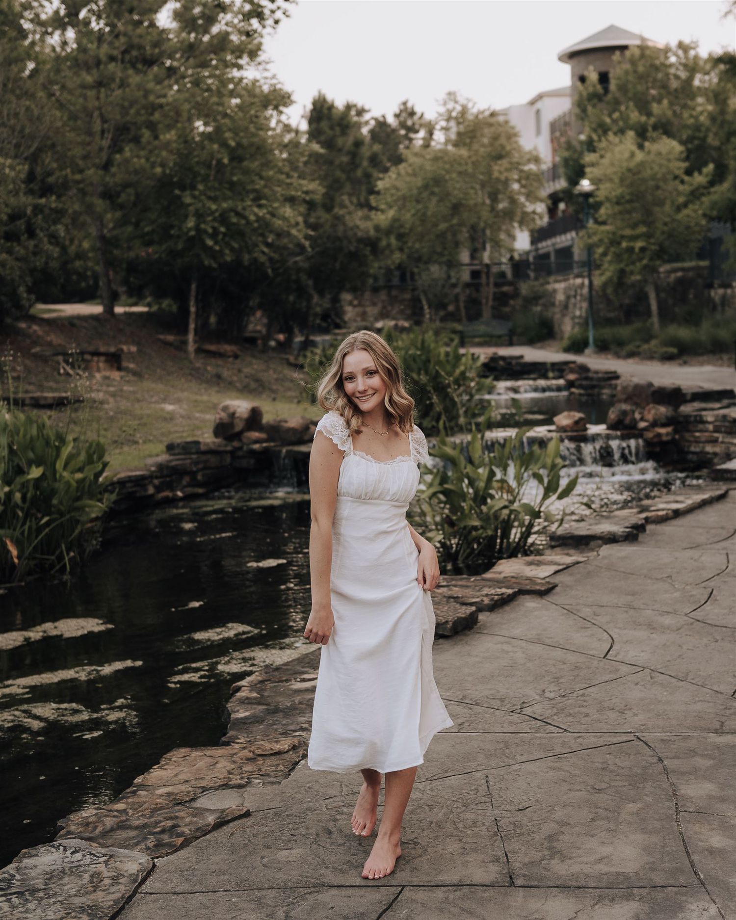 Someone in a white dress poses on stone steps near a decorative pond and waterfall.