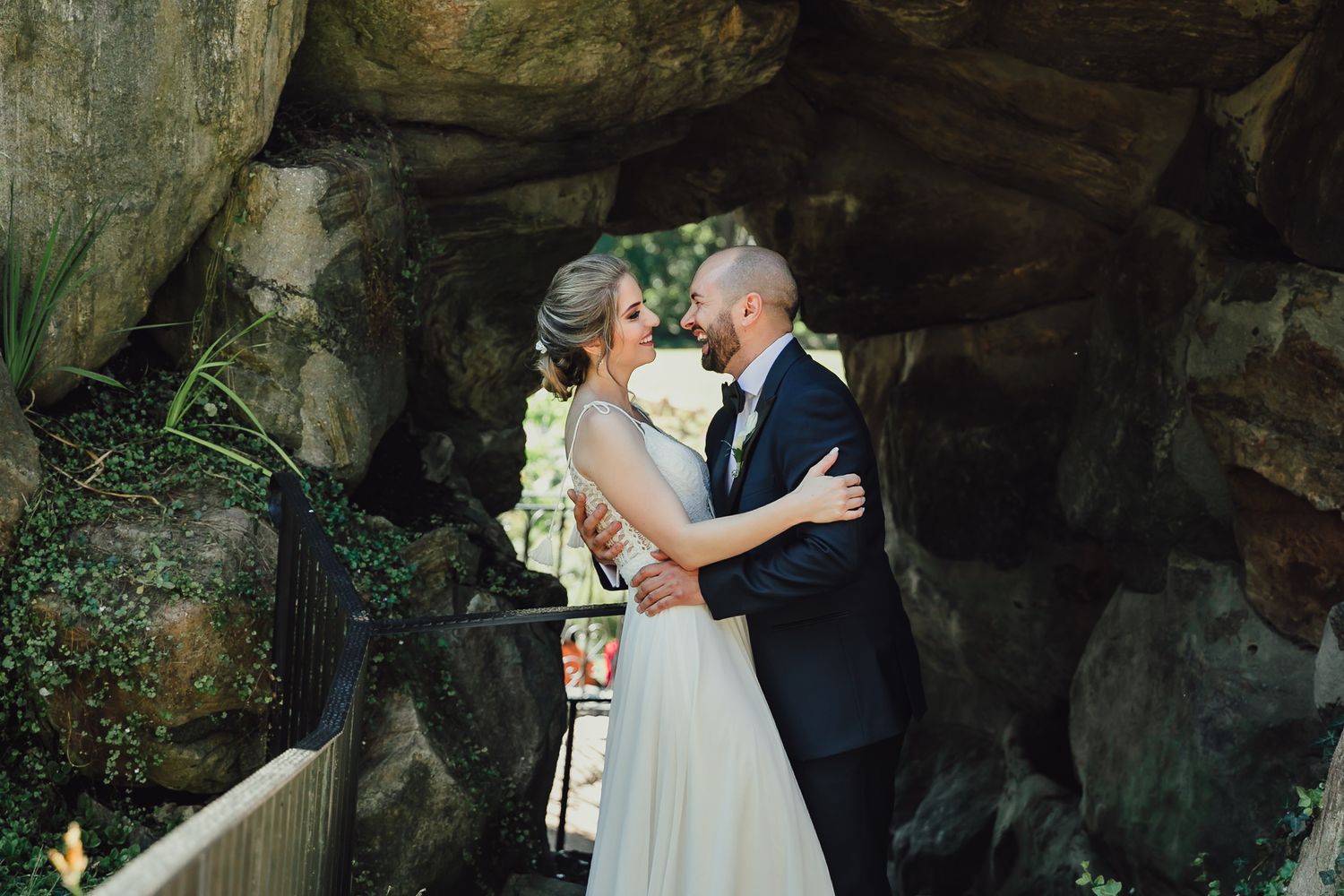 Romantic bride and groom share intimate moment near rustic stone grotto at outdoor wedding.