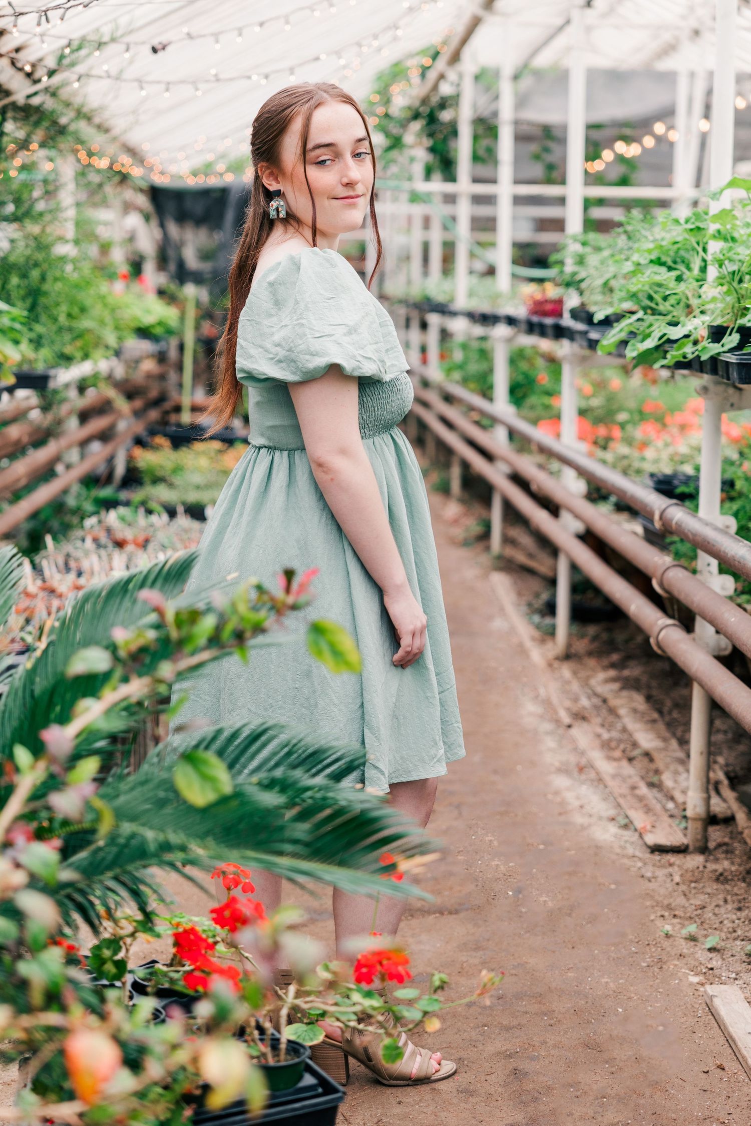 Person in vintage-style mint dress poses among greenhouse plants and red flowers.