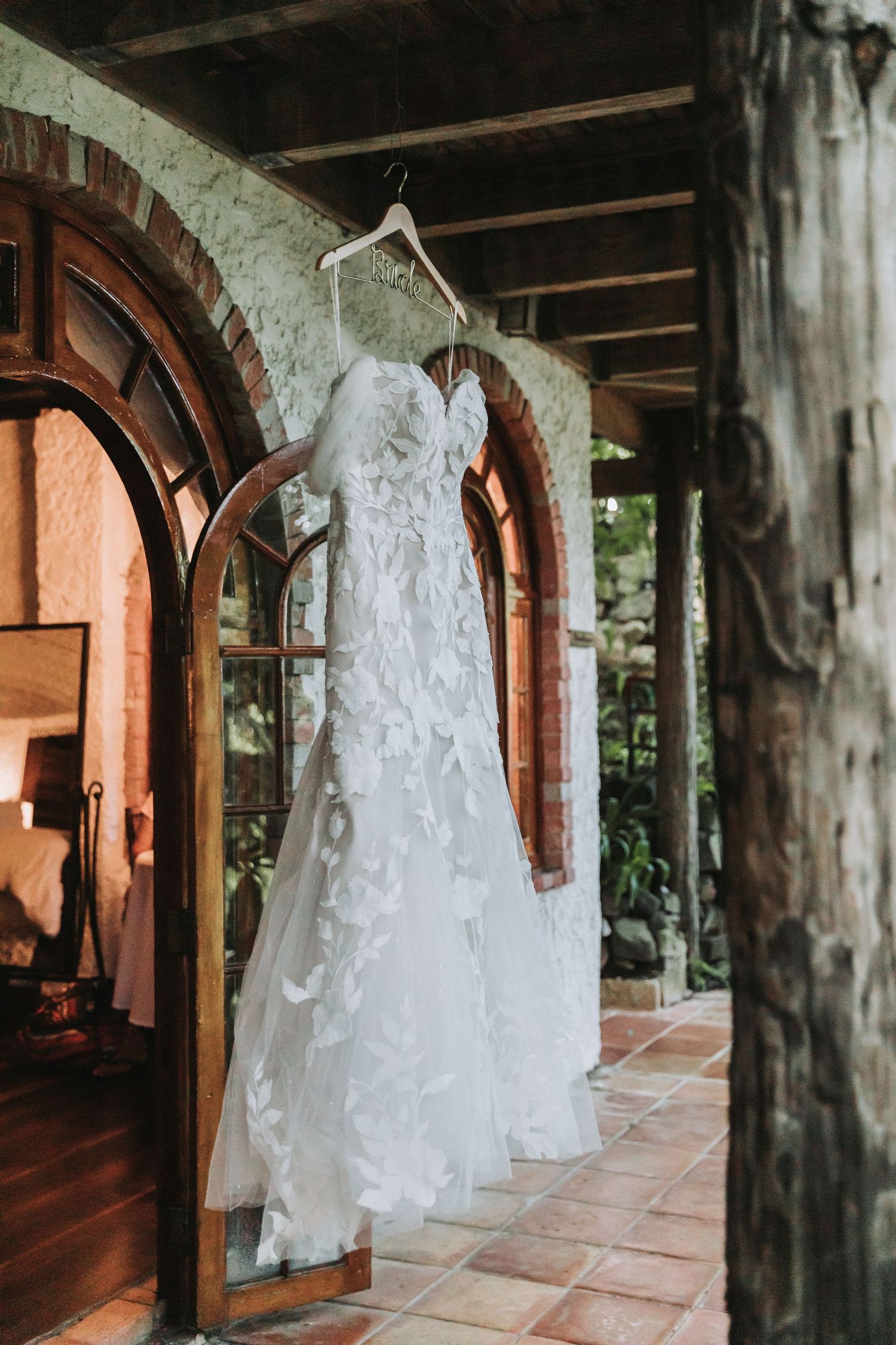 A delicate white lace wedding dress hangs in an arched doorway of a rustic building.