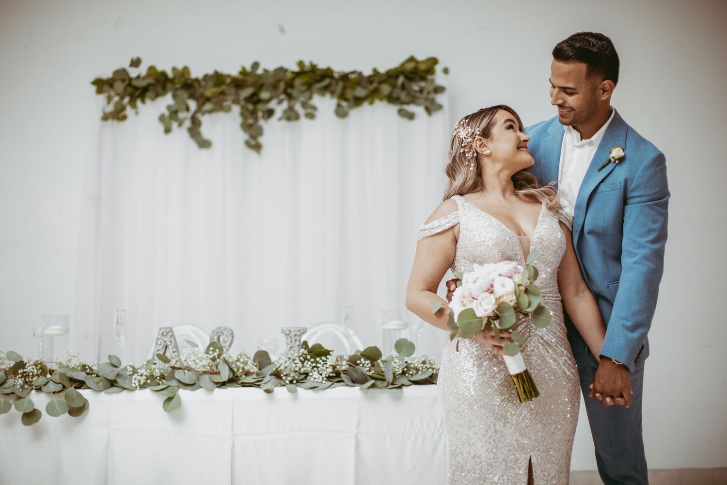 Wedding couple sharing tender moment against white backdrop with greenery decorations.