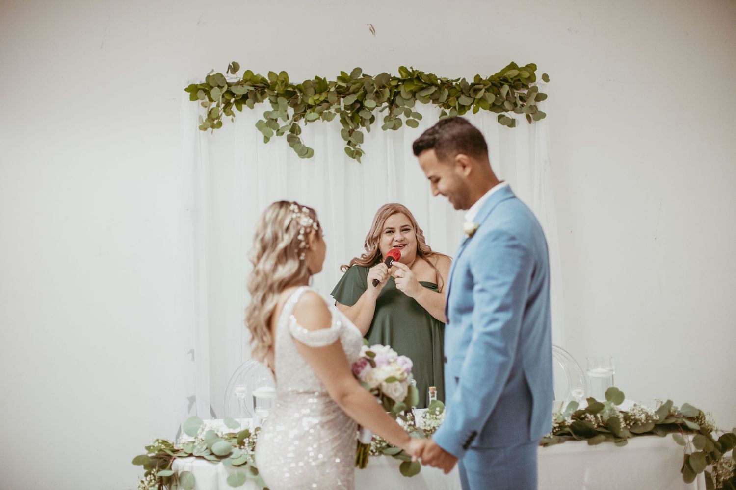 Wedding ceremony with couple and officiant under green floral arch against white backdrop.