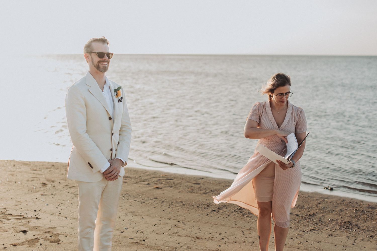 A couple stands on a beach at sunset wearing light colored formal attire.