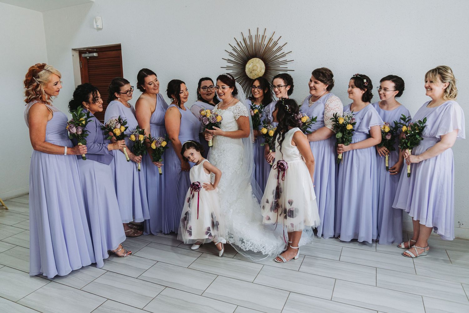 Wedding party lined up indoors wearing lavender bridesmaid dresses and light-colored suits.