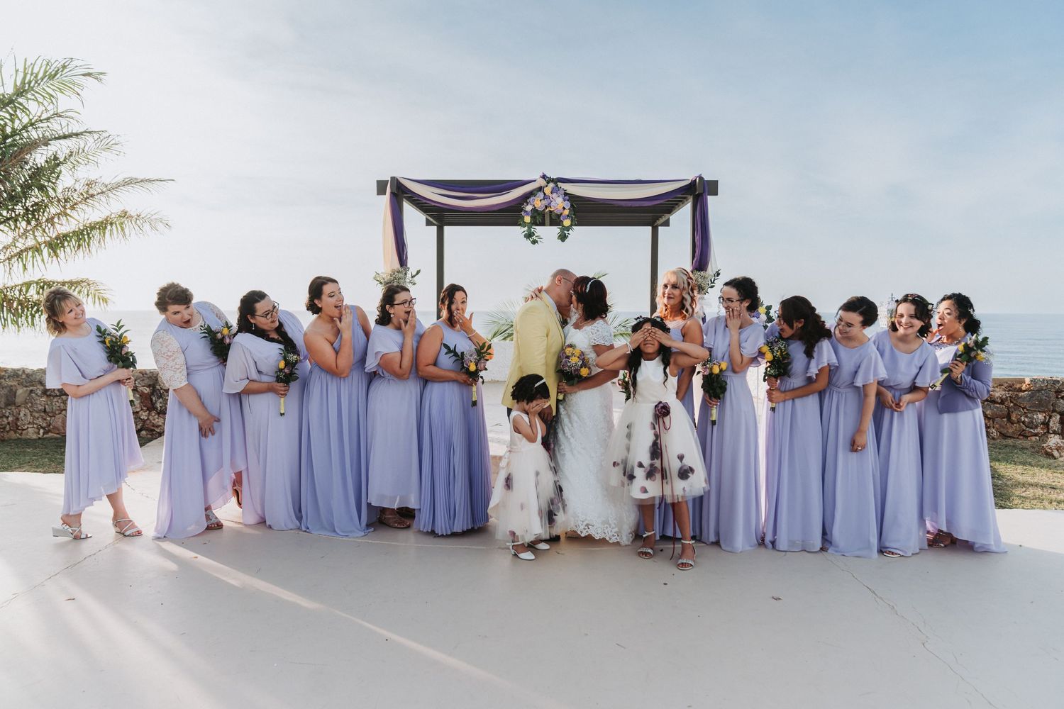 Wedding party gathered under purple-draped arch at outdoor beach ceremony location.