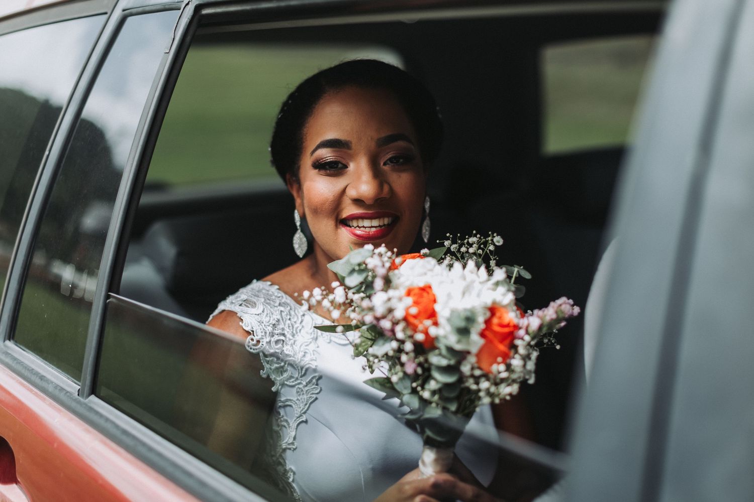 Bride smiling through car window while holding wedding bouquet with orange flowers.