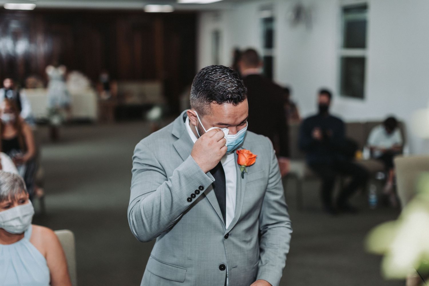 Emotional groom in grey suit and face mask wipes tear during ceremony.