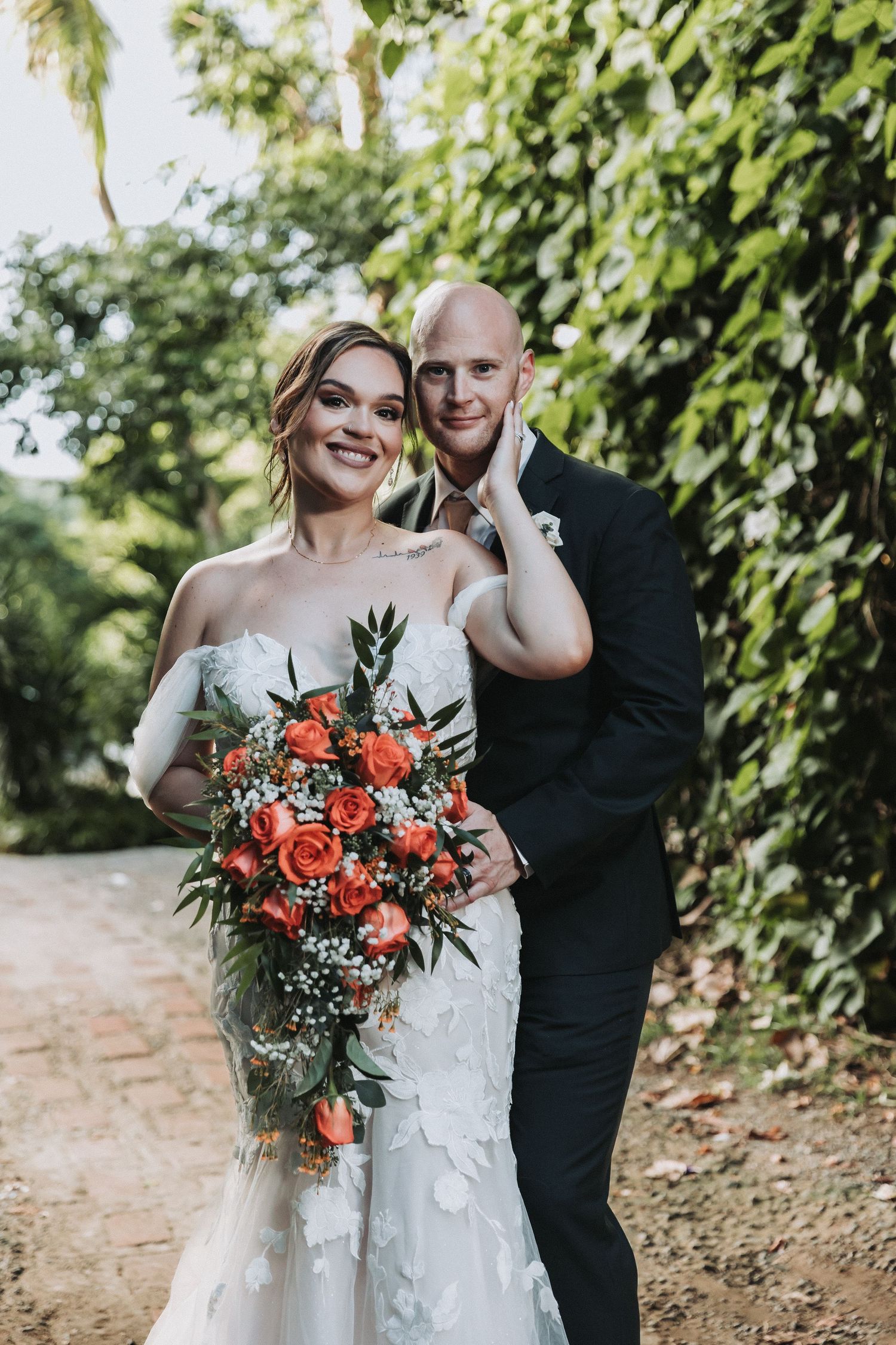 A newlywed couple poses on a garden path with a vibrant coral rose bouquet.