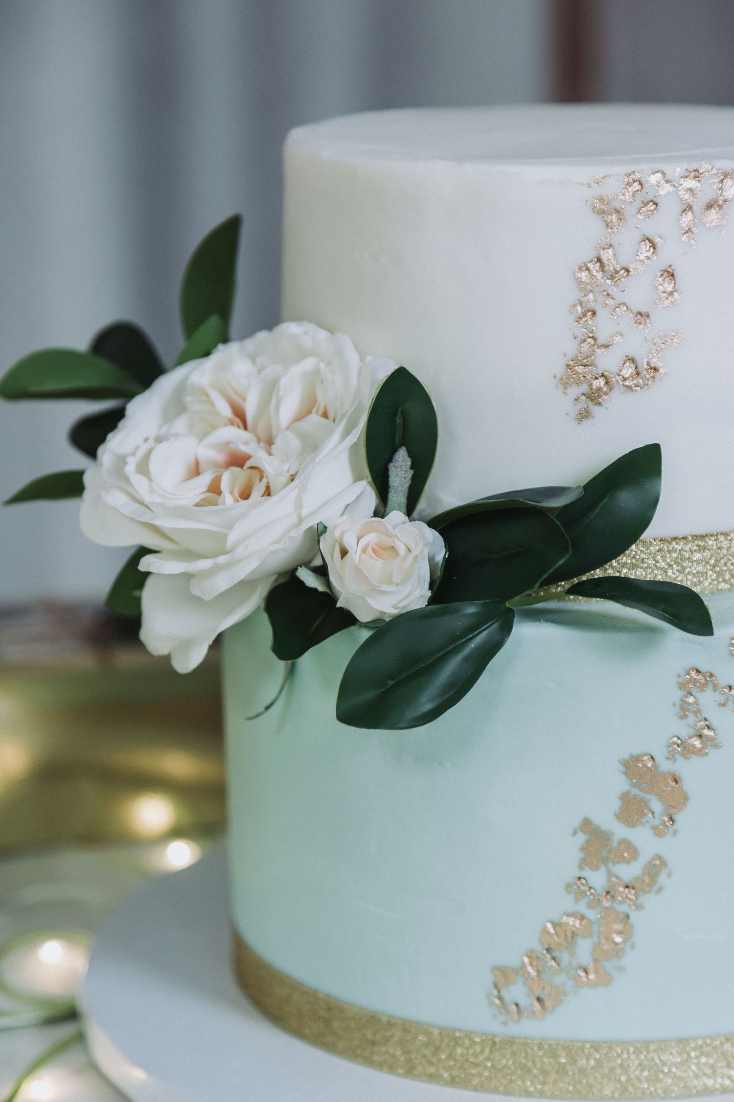 An elegant wedding cake decorated with white roses and gold accents on a mint green tier.