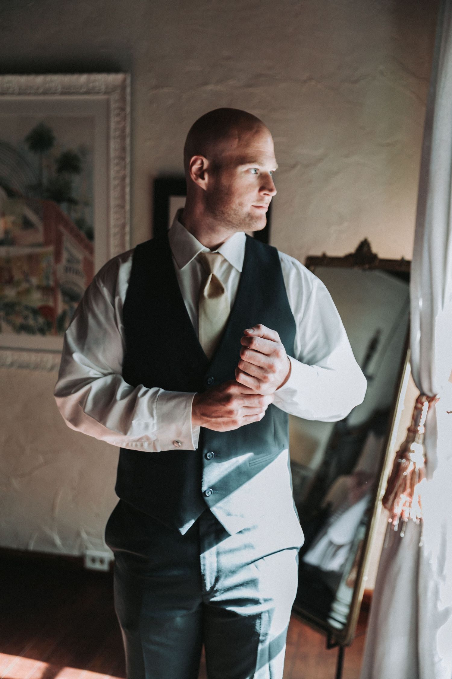 A groom in formal attire adjusts his tie while getting ready in a sunlit room.