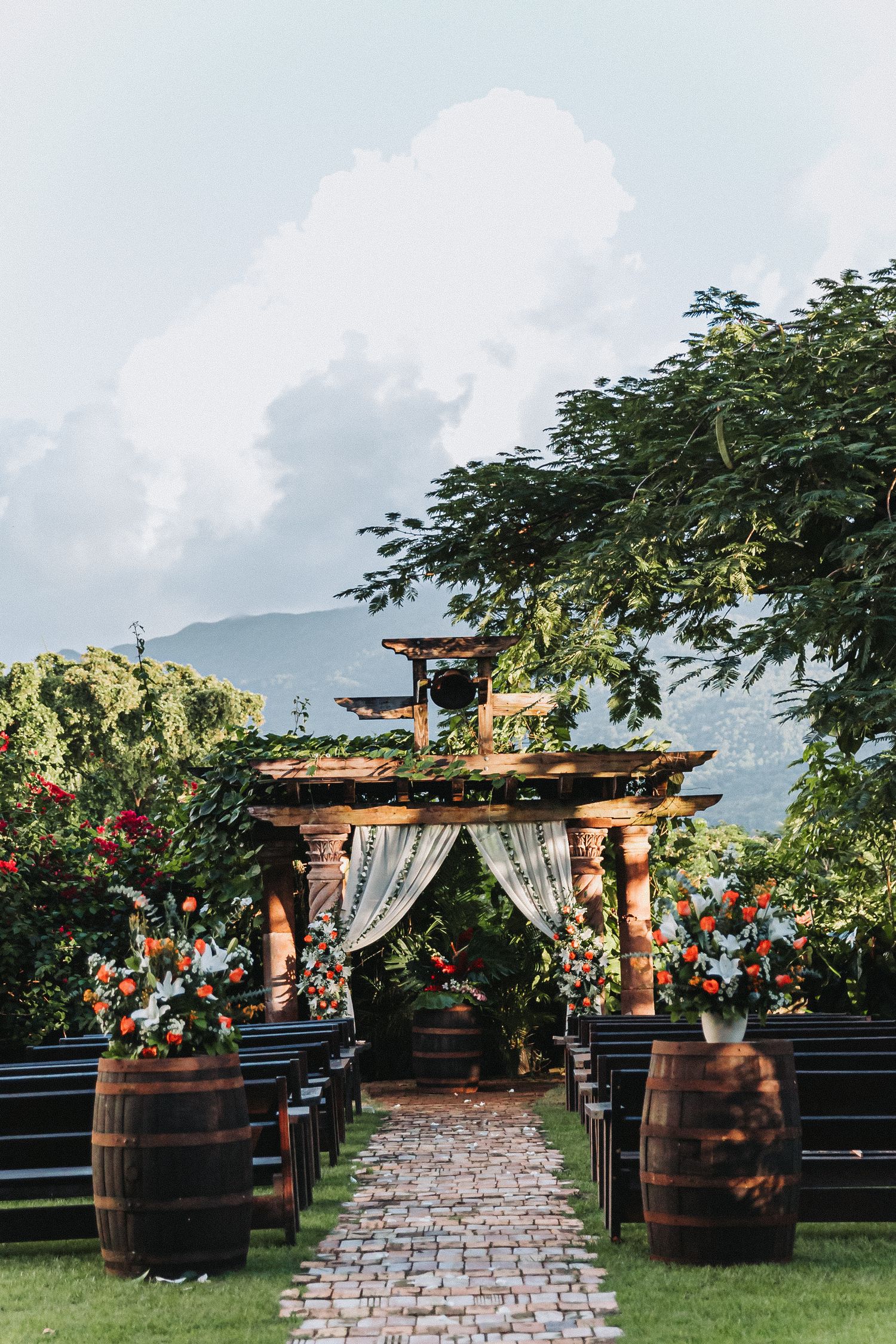 Romantic outdoor wedding ceremony setup with wooden arch, flowing curtains, wine barrels and mountain scenery in background.