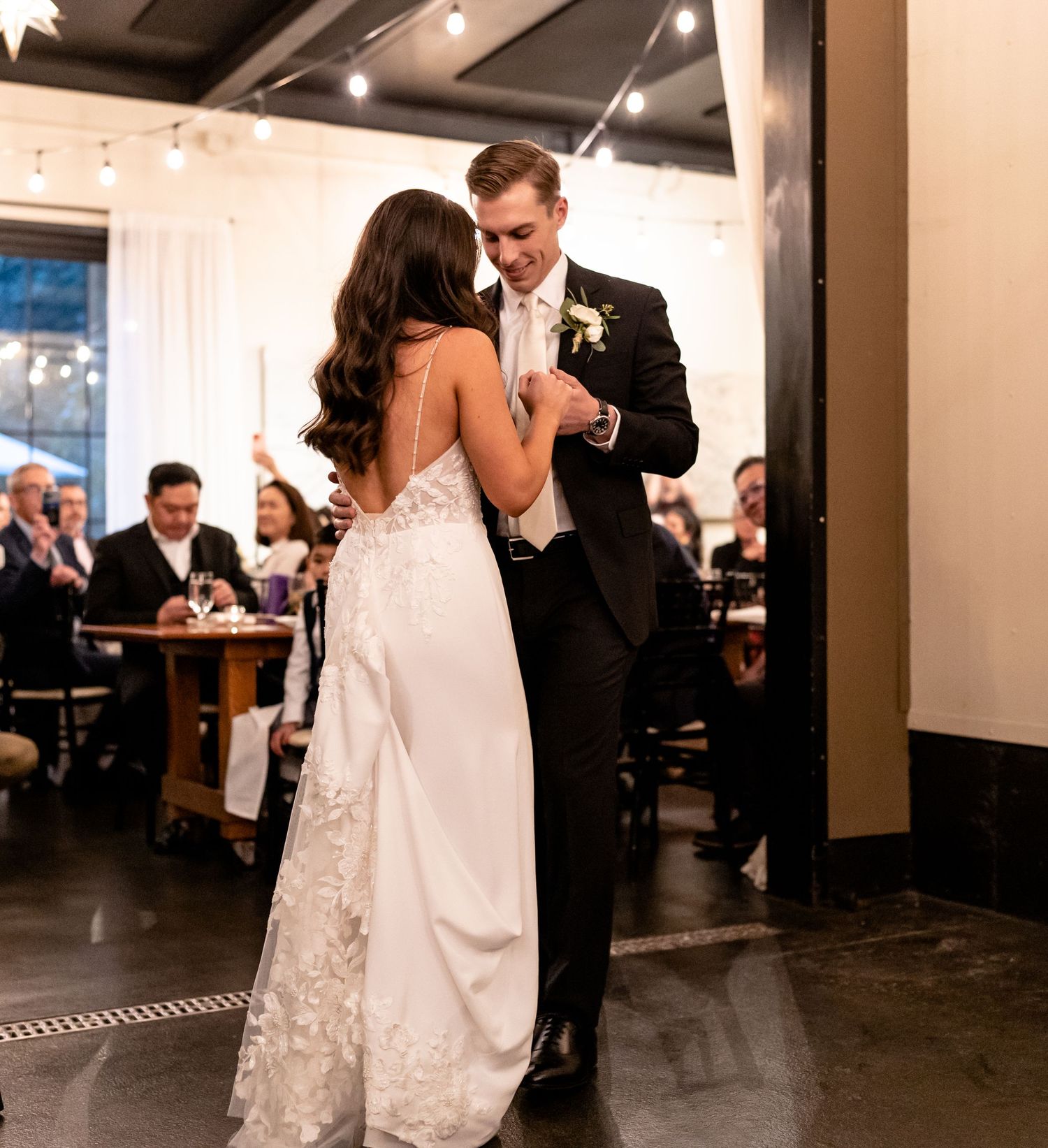Couple shares their first dance at a wedding reception with guests seated at tables in the background.