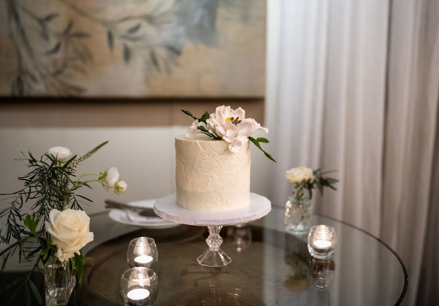 Simple elegant wedding cake with pink floral decoration displayed on glass stand with candlelight.