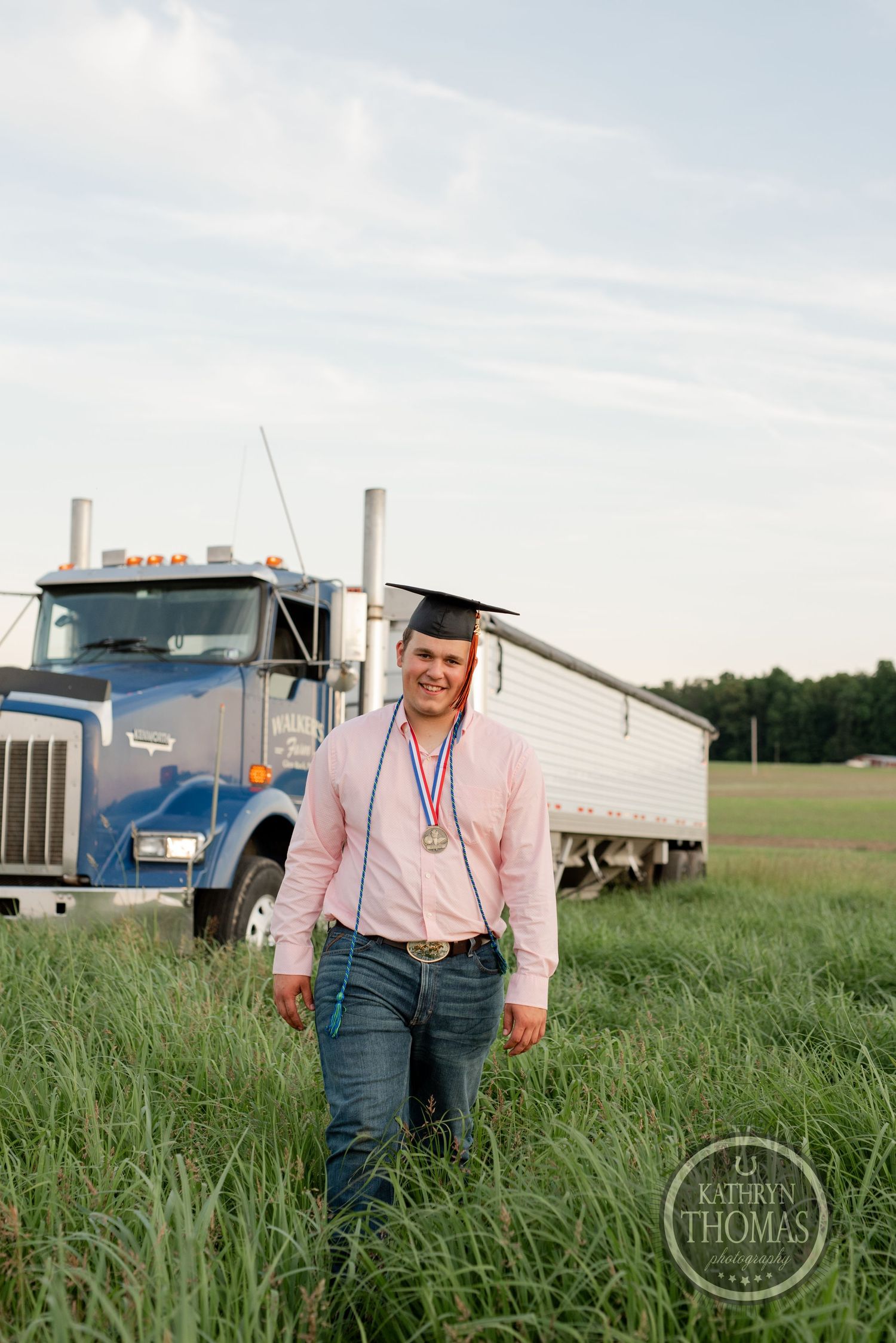 Graduate in pink shirt and graduation cap standing in field with semi truck in background.