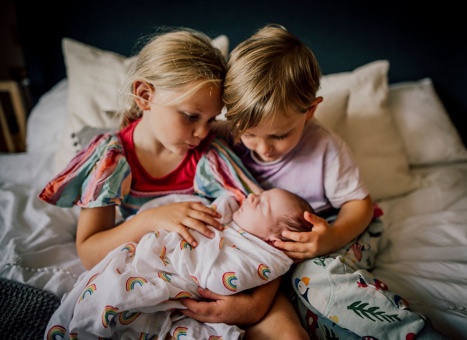 Two children sitting on a bed tenderly holding and looking at a newborn baby wrapped in blankets. Worcestershire Wedding photographer.