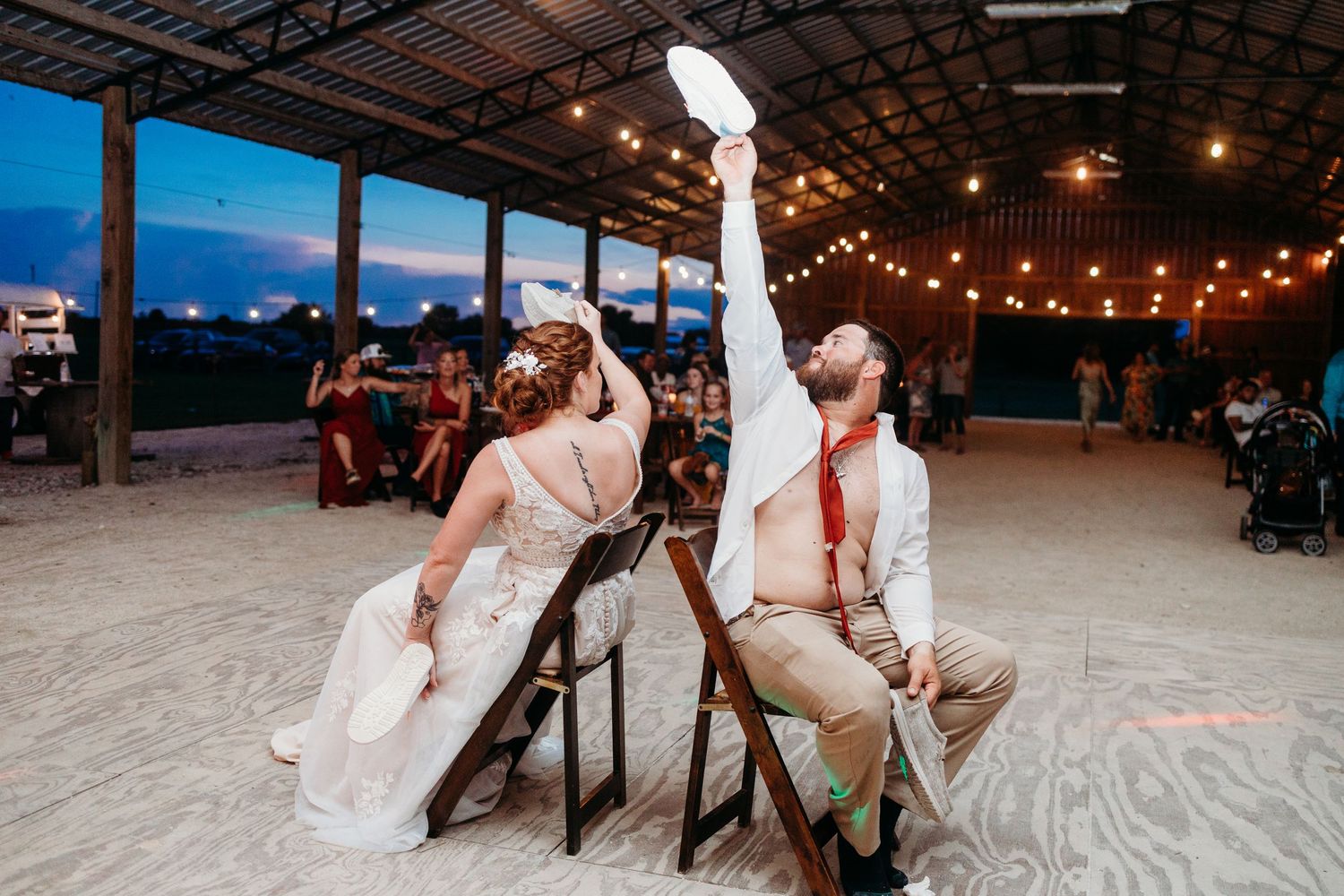 A couple plays a fun wedding game sitting on chairs in an open-air venue with string lights.