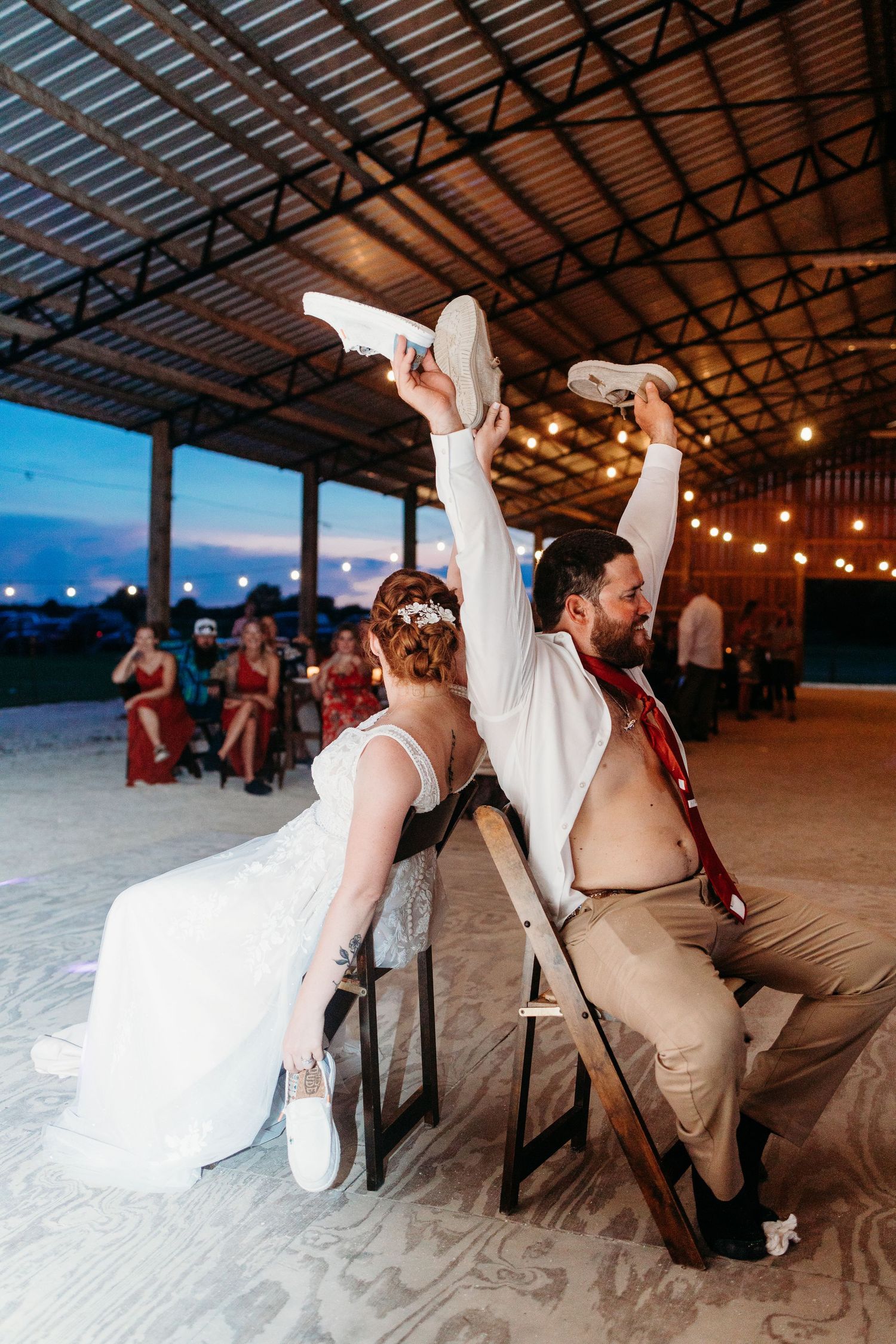 A playful wedding couple participates in a reception game on chairs at an outdoor covered venue.