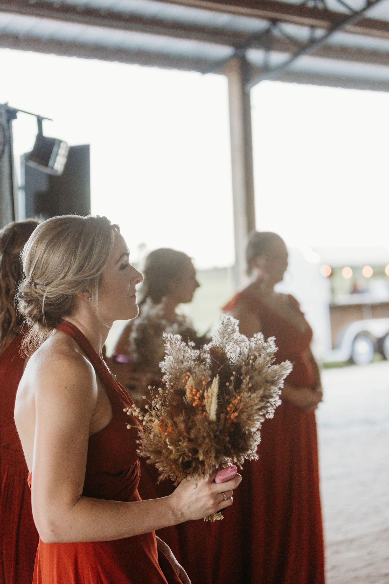 Bridesmaids in burgundy dresses hold dried flower bouquets at an outdoor wedding venue.