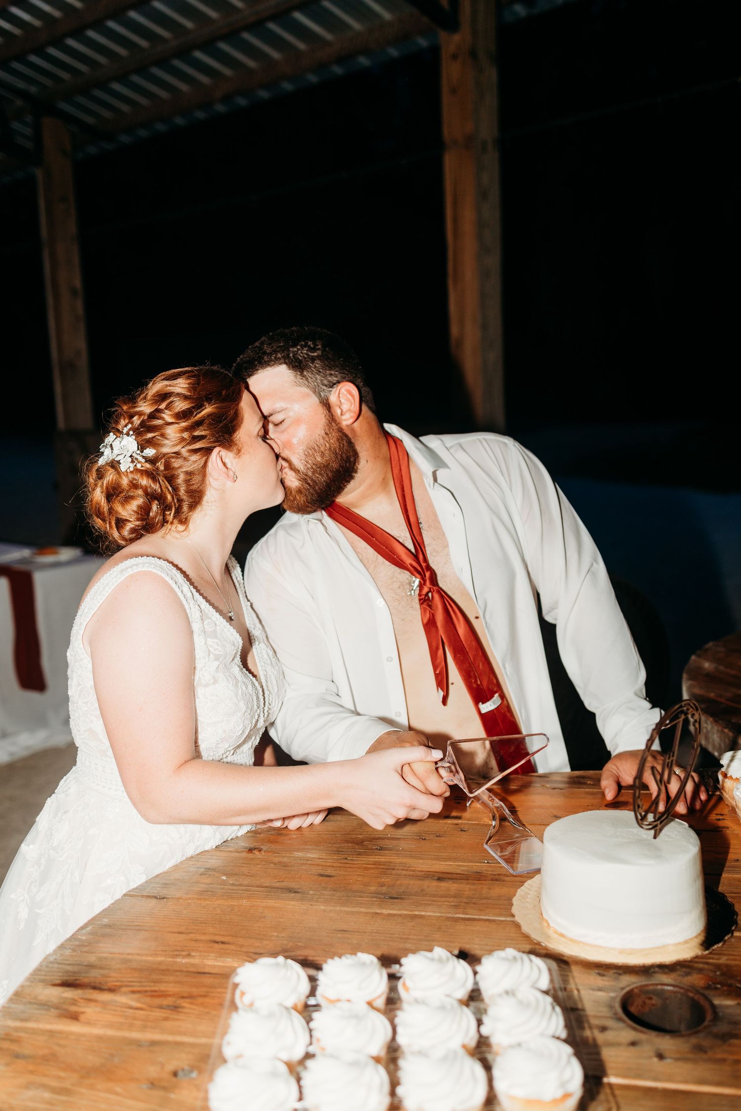 A sweet moment shared between newlyweds at their wedding cake table in a rustic barn setting.