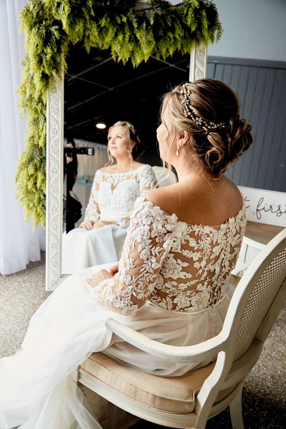 Elegant bride in lace gown sits at vintage vanity mirror with cascading greenery overhead.