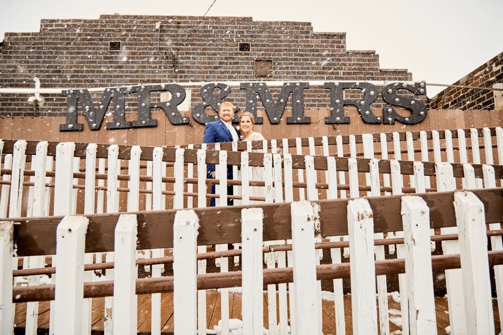 A couple poses playfully by a white picket fence with 'Mr & Mrs' signage above.
