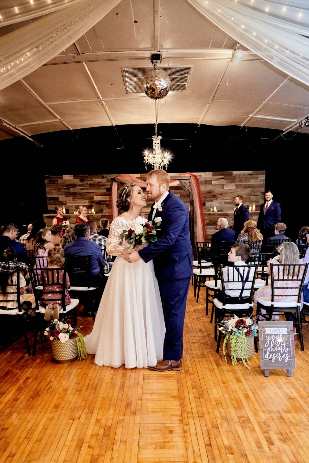 Wedding ceremony in rustic venue with wooden backdrop, chandeliers, and white draping.