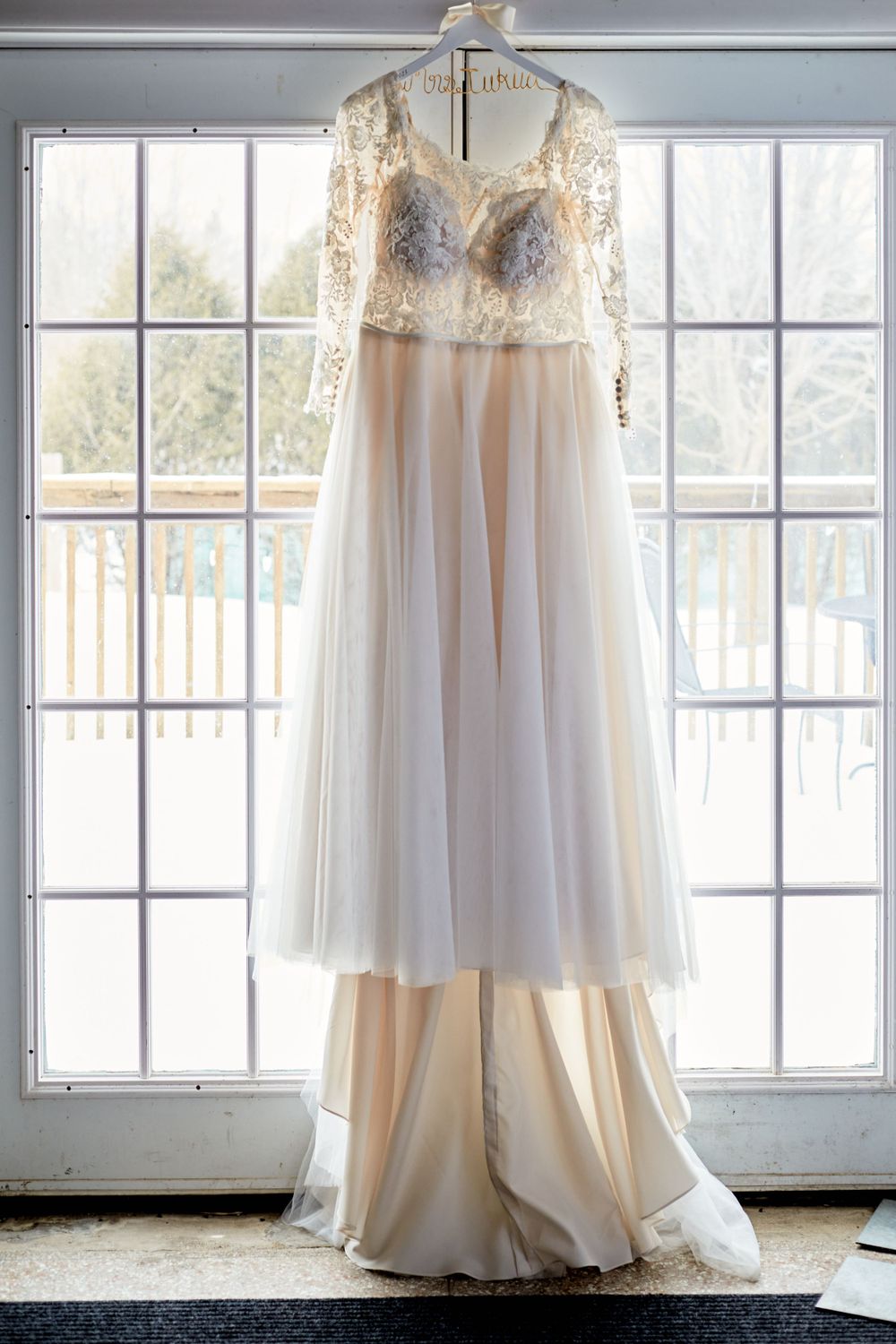 Delicate lace wedding dress hangs in window displaying intricate bodice details.