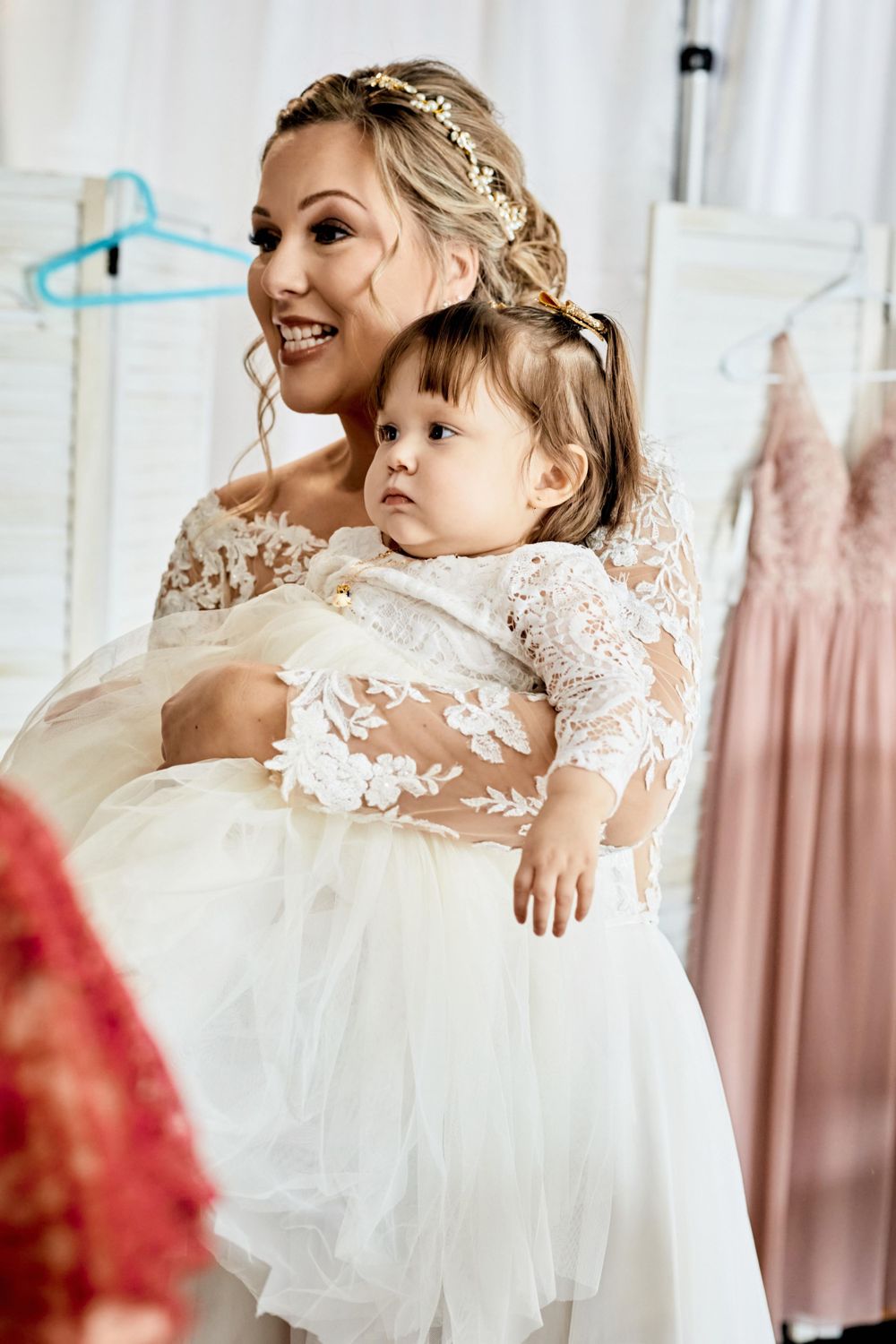 A bride in a lace wedding gown holds a small child while getting ready.