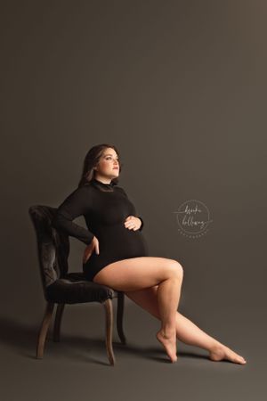 Artistic portrait of figure in black outfit posing on vintage chair against dark backdrop.