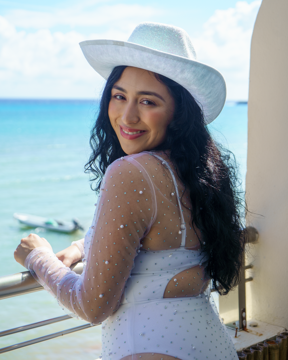 Woman in white dress and hat poses against beautiful turquoise ocean backdrop.