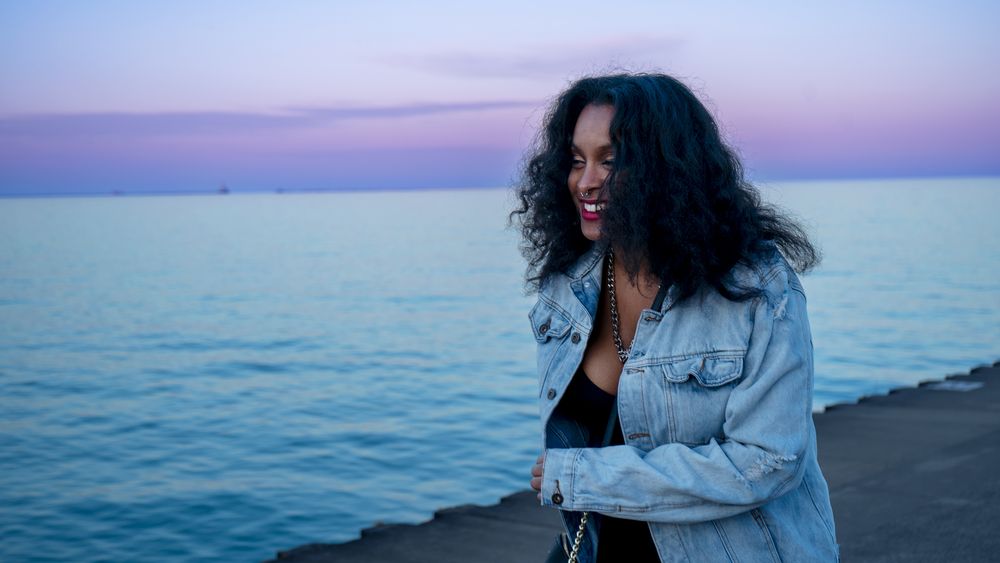 Woman with curly hair in denim jacket enjoys sunset by the ocean.
