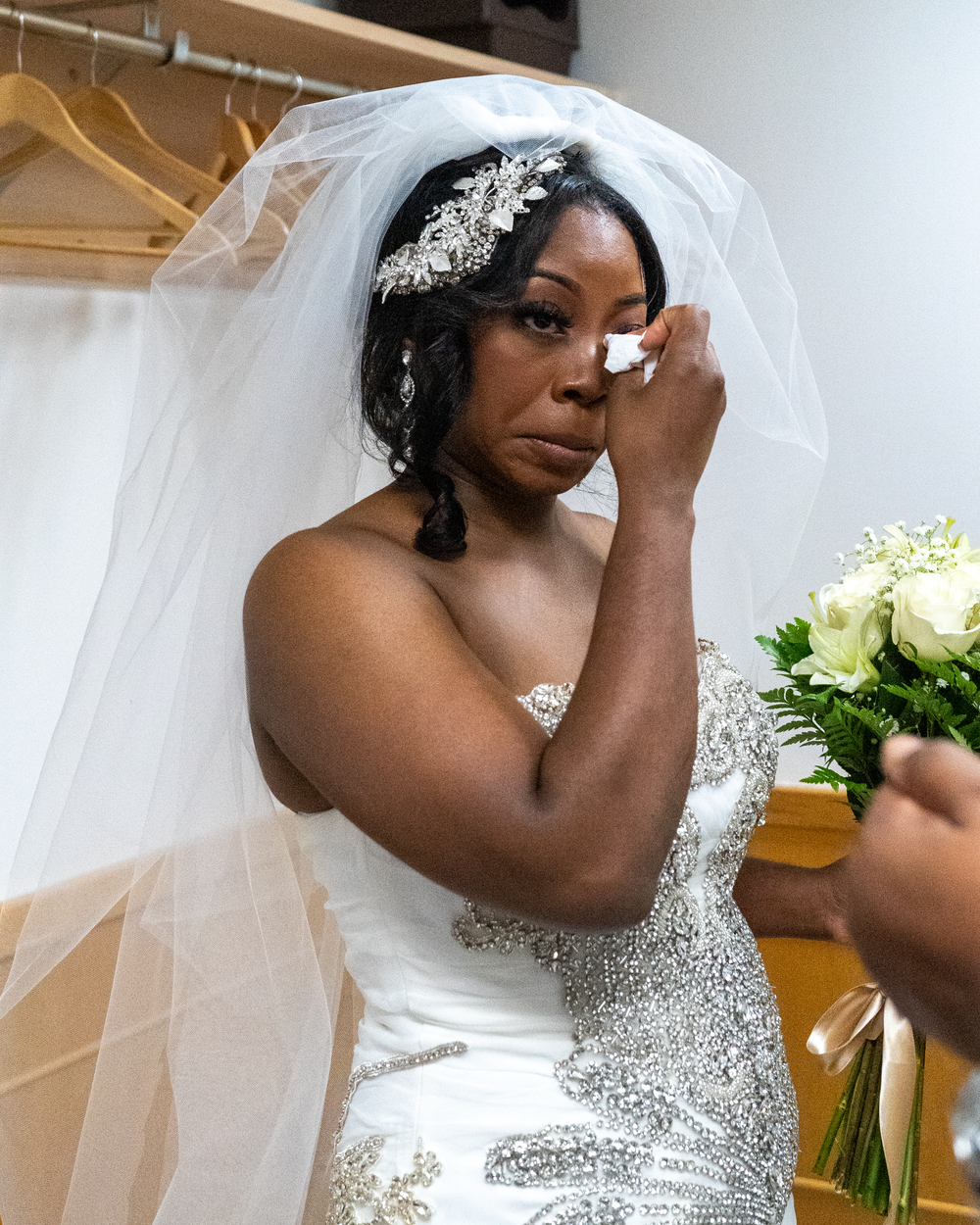 Emotional bride in sparkling white wedding dress wipes tear while holding bouquet.