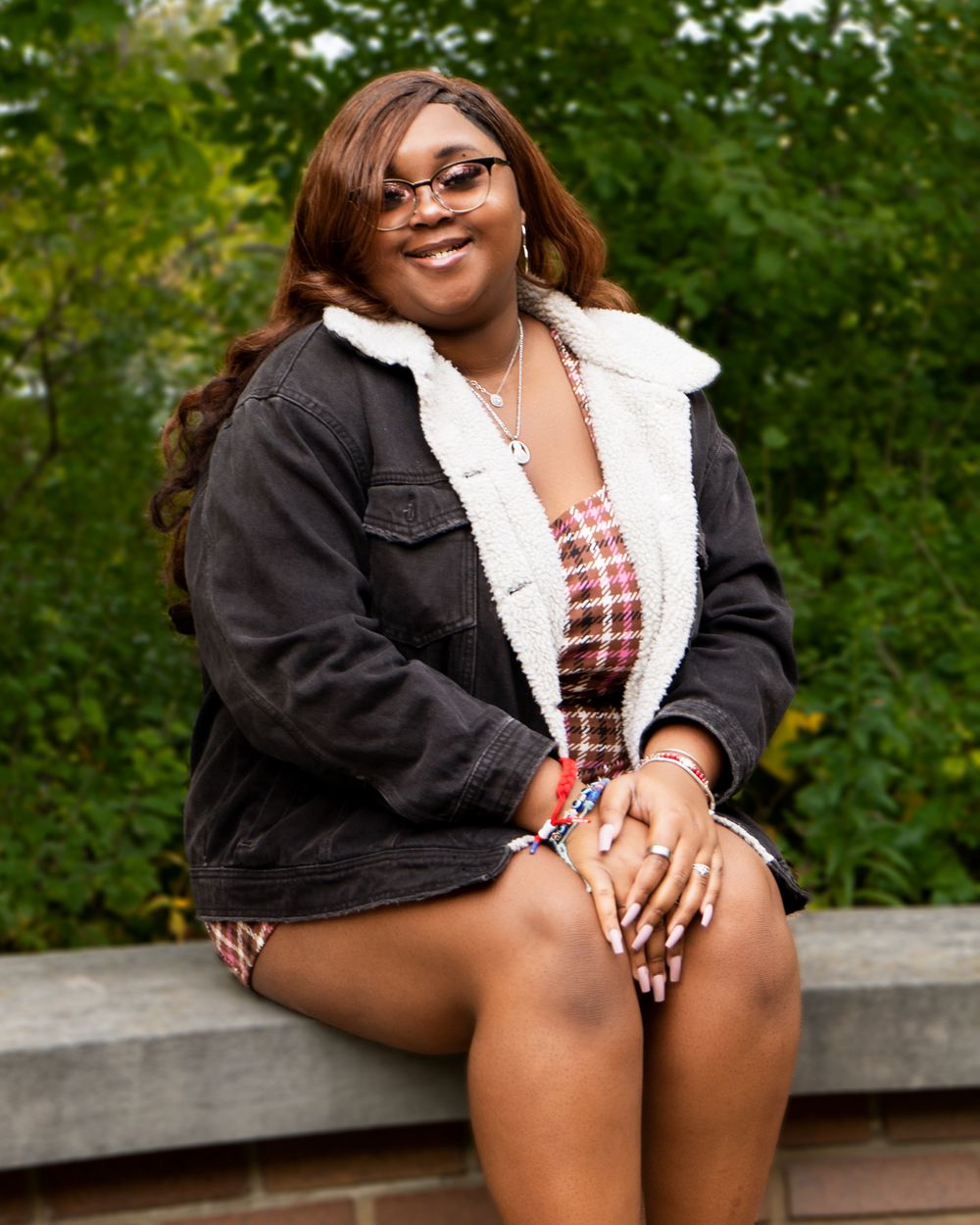 Smiling woman in denim jacket and plaid top sits outdoors on wooden bench.