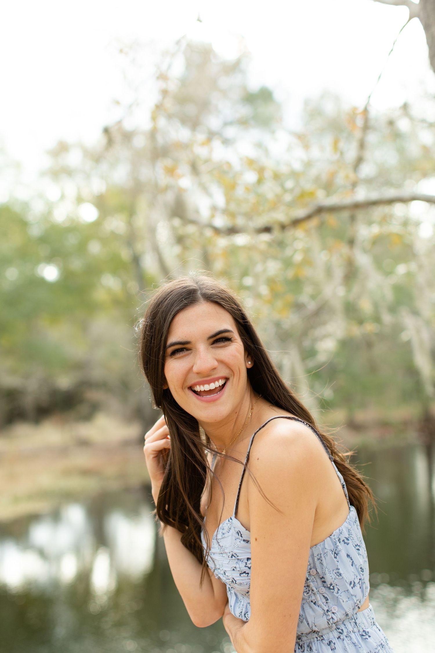 Woman in blue dress laughs joyfully while posing near a pond with trees in background.