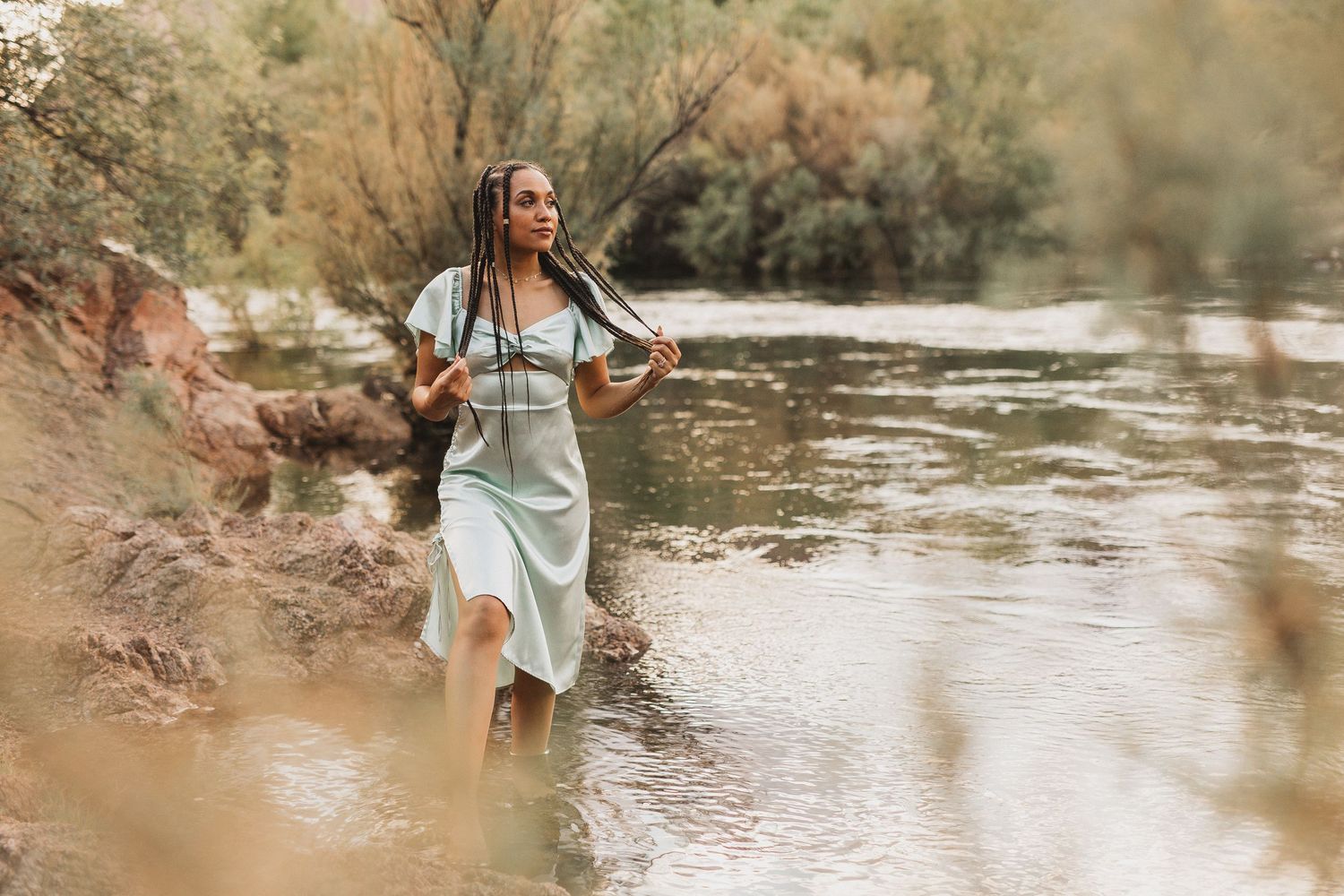 Person in flowing dress standing joyfully by riverside in natural setting.