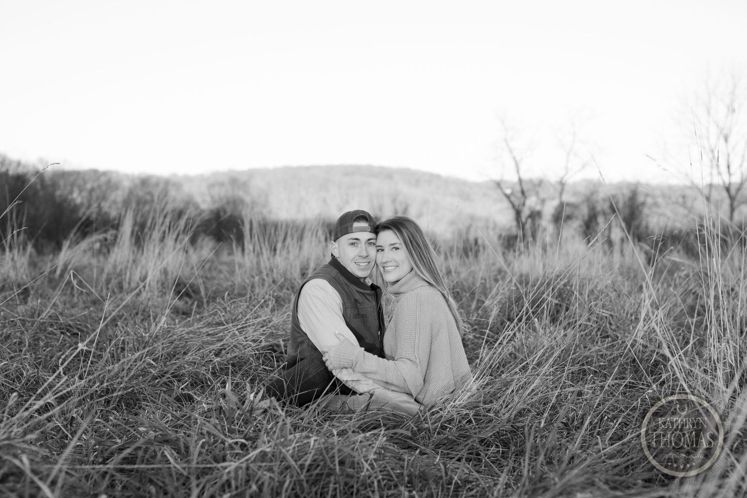 A black and white portrait of a couple cuddling in long grass with rolling hills in the background.
