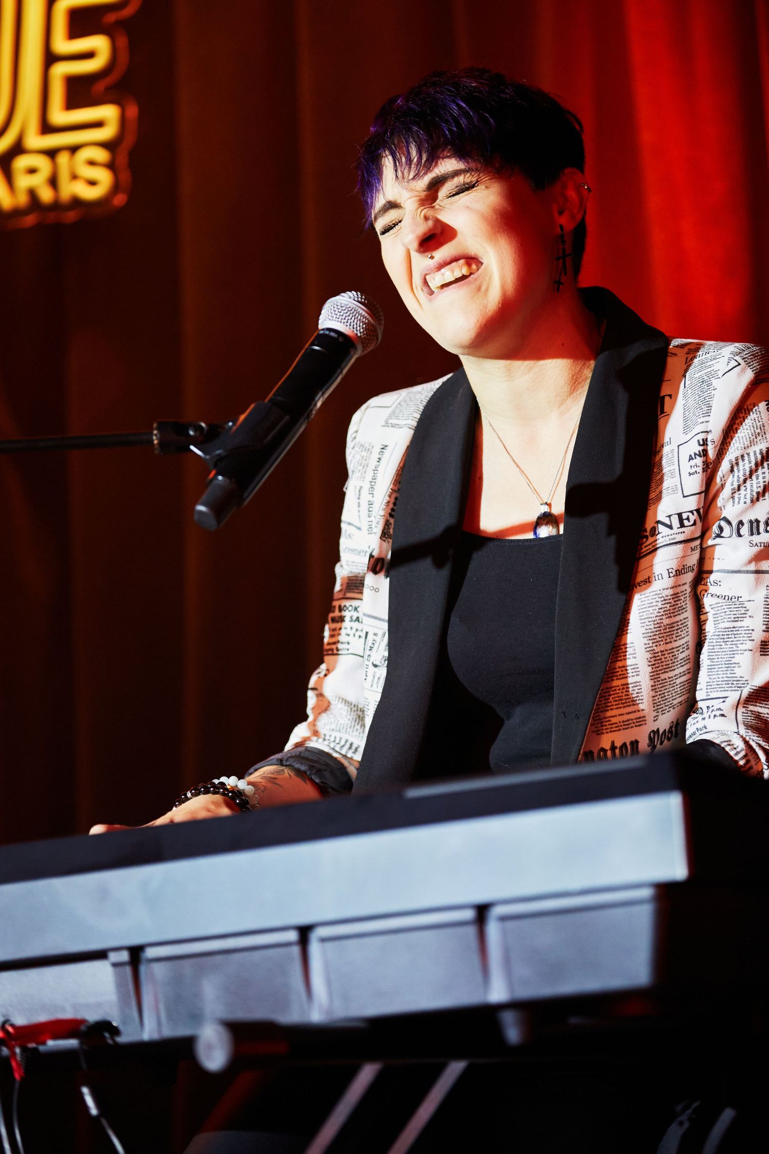 Performer singing and playing keyboard at a concert venue with red lighting and neon signage.