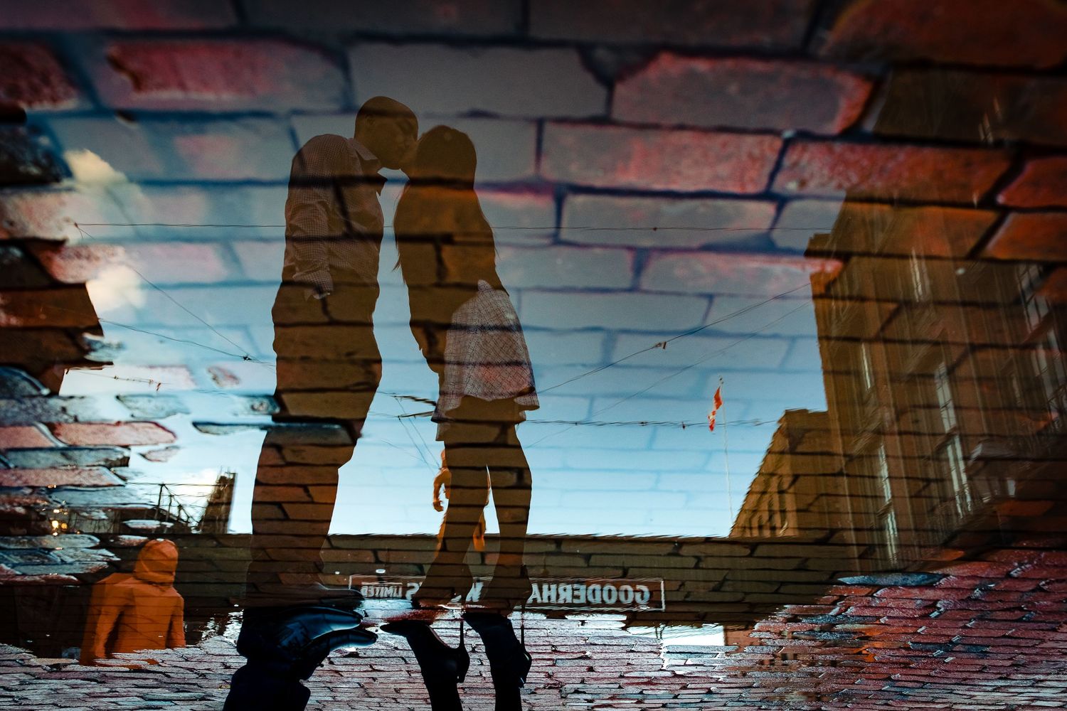 Silhouetted couple reflected in puddle against brick wall and blue sky backdrop.