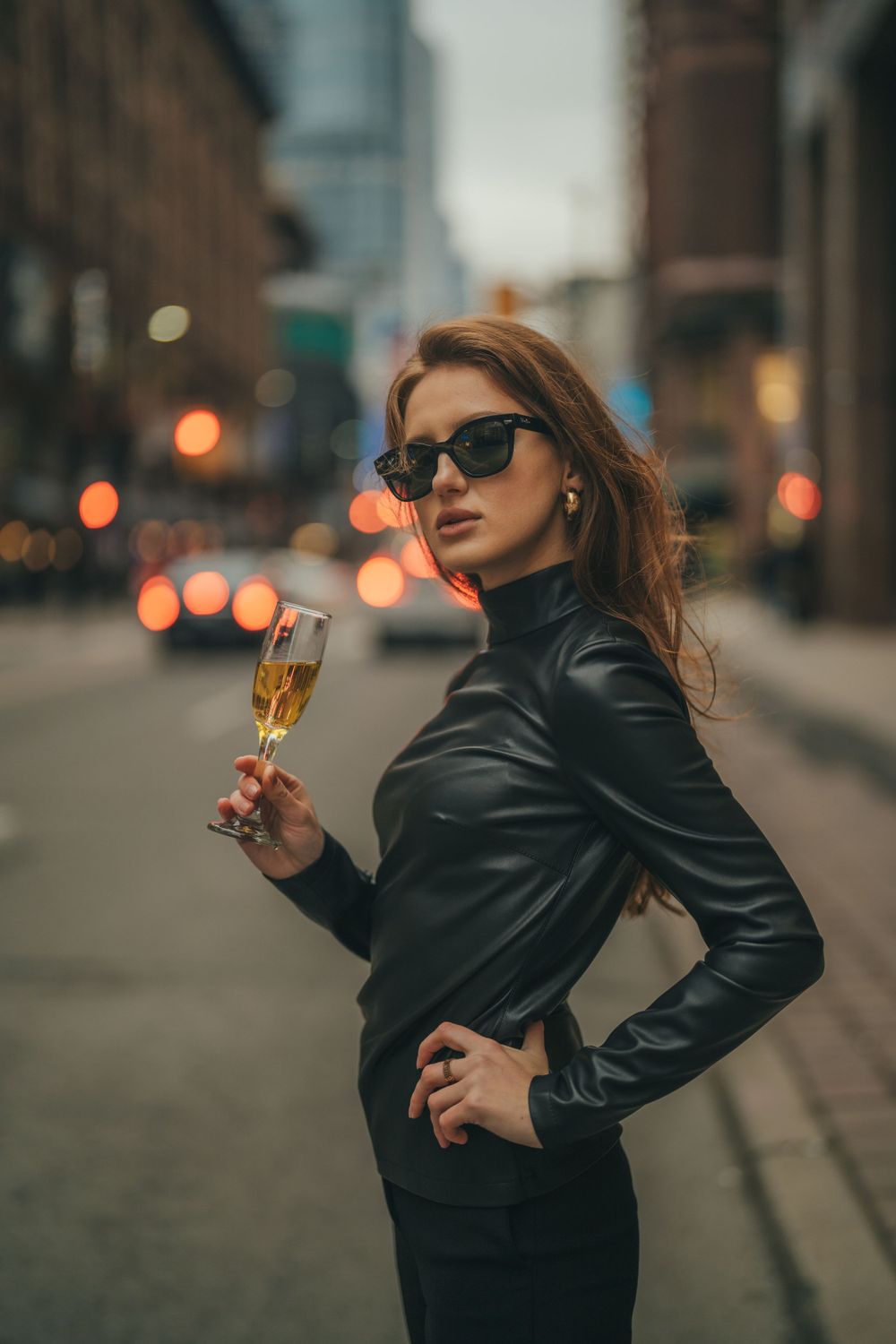 Stylish person in leather jacket and sunglasses holds champagne glass on city street at dusk with bokeh lights.