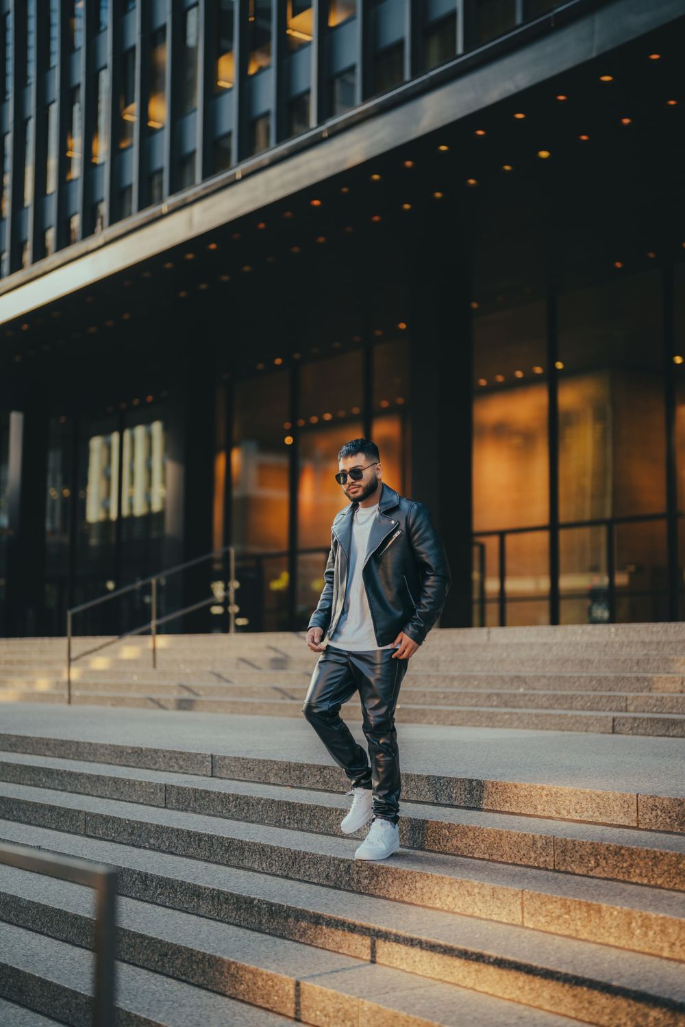 Fashionable person in leather jacket and sunglasses poses confidently on modern building steps at dusk with warm lighting.