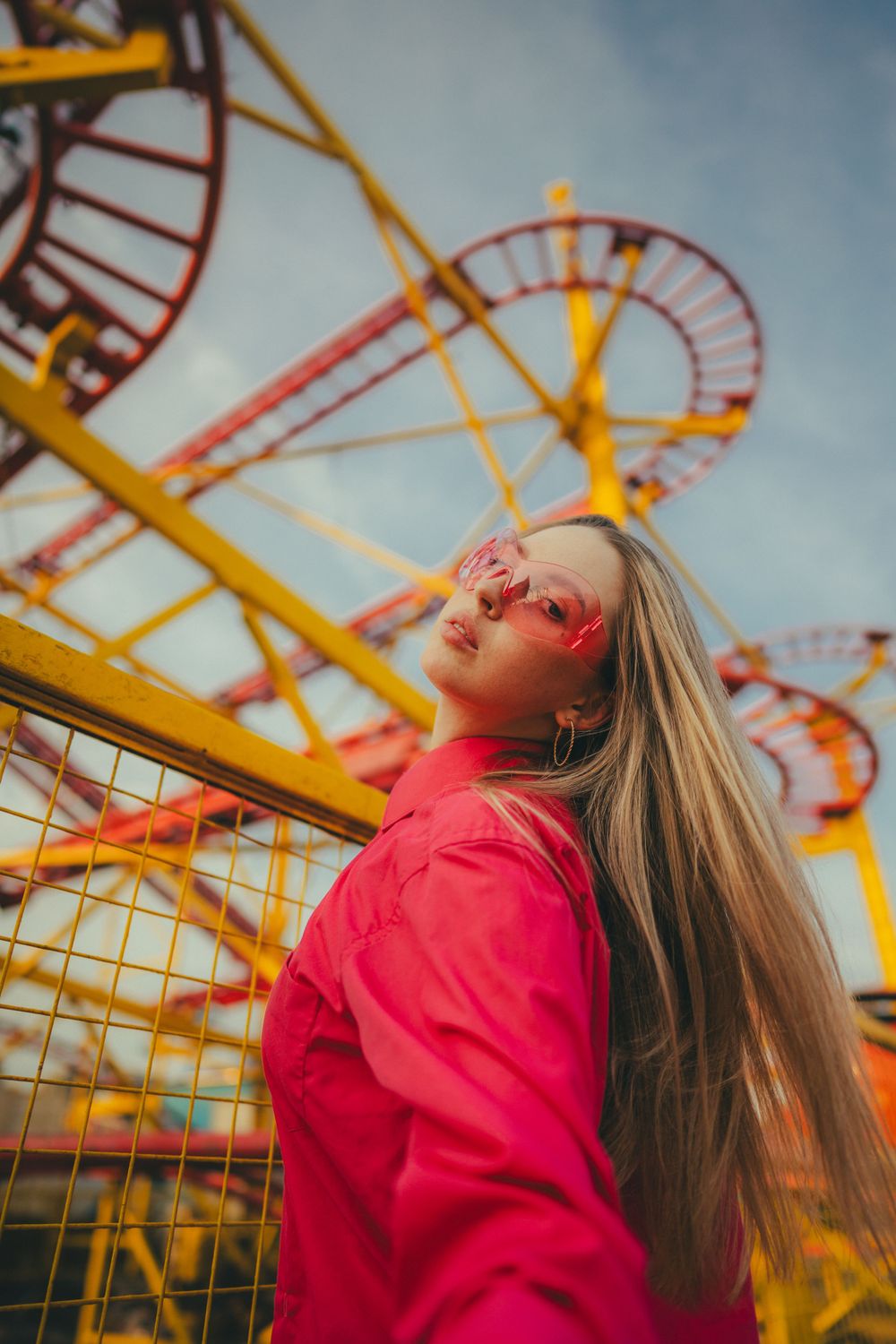 A person in a bright coral jacket poses dramatically against a vibrant yellow and red roller coaster background at sunset.
