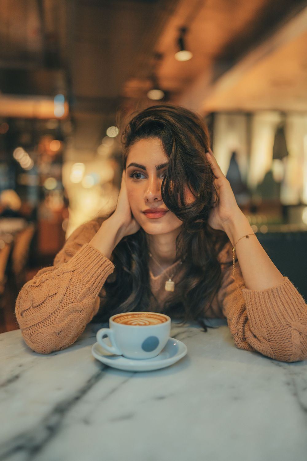 Person in cozy sweater relaxing at cafe with latte art coffee on marble table in warm ambient lighting.