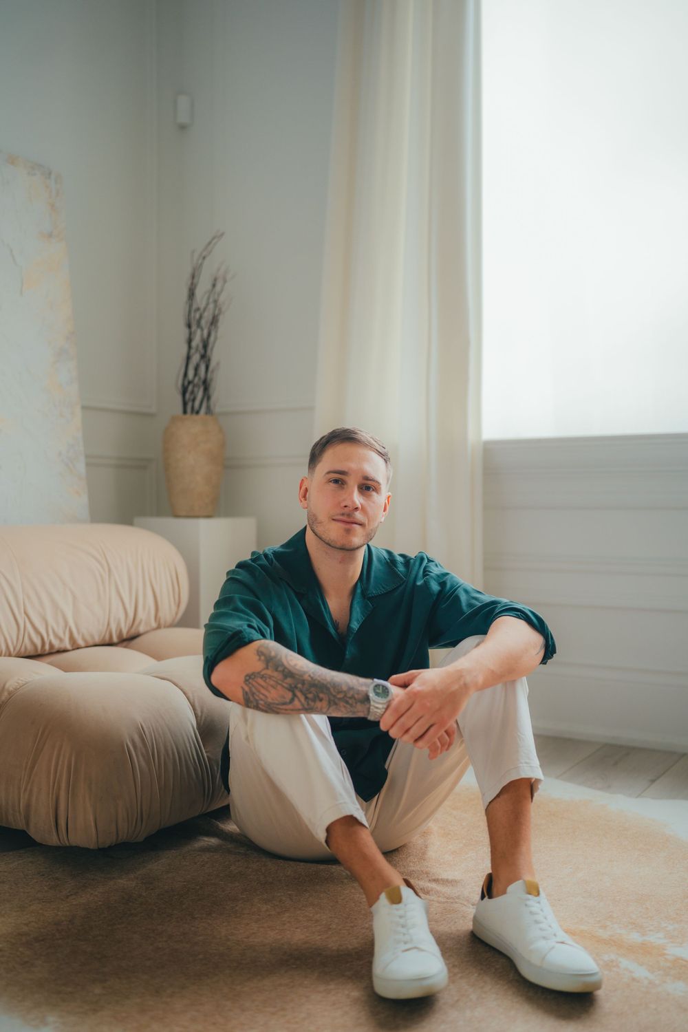 Someone poses casually on a carpeted floor wearing a green button-up shirt, white pants and white sneakers in a minimalist room.