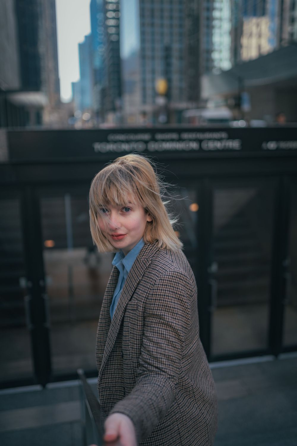 Person in a plaid blazer and blue shirt stands on an urban street with skyscrapers in the background at dusk.