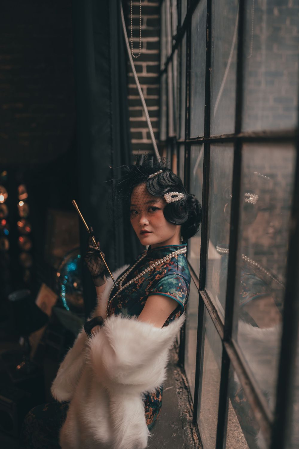 A person in a traditional blue Chinese qipao dress poses by a window with moody lighting and vintage ambiance.