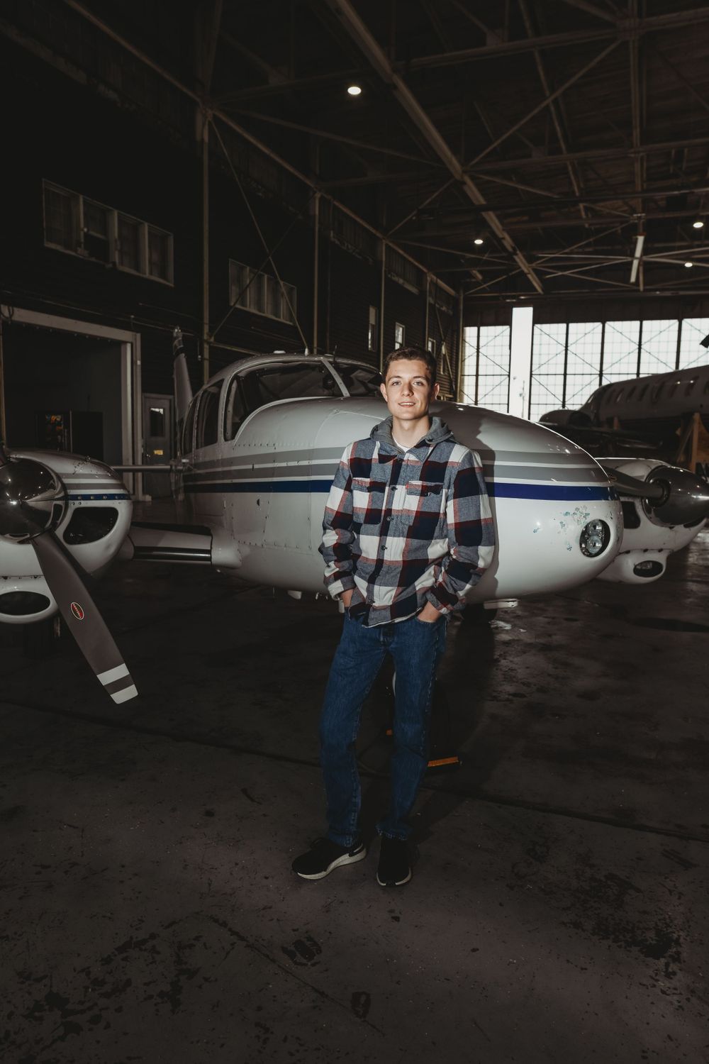 Person in plaid shirt stands in aircraft hangar with planes in background.