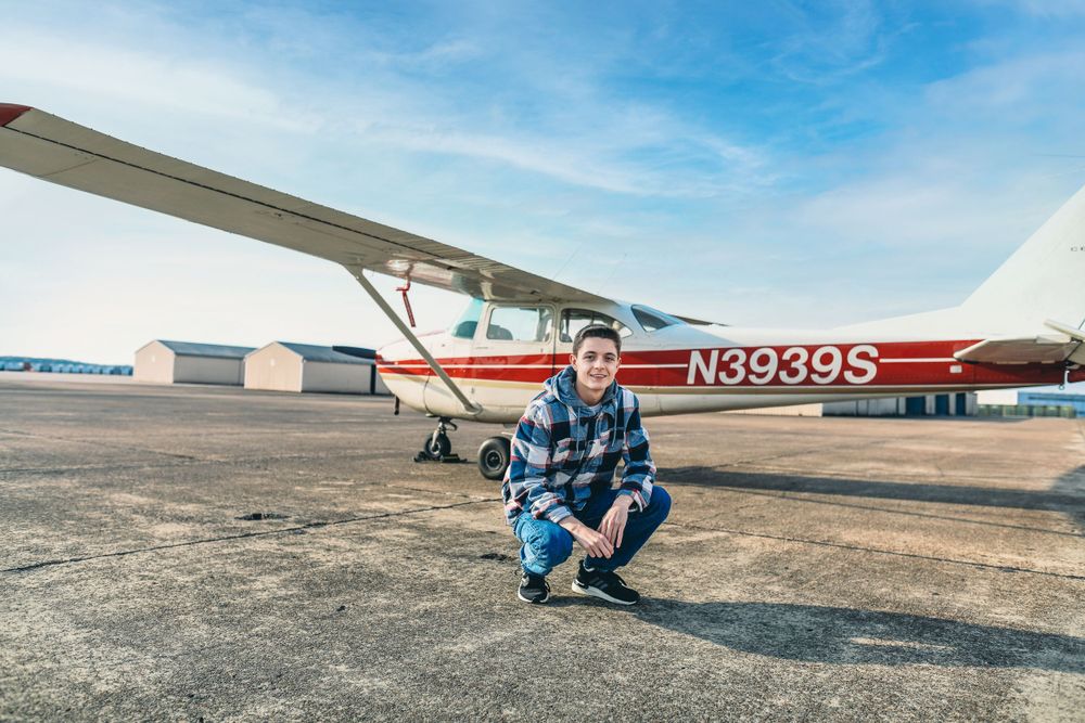 Person stands on airfield with small aircraft and blue sky in background.