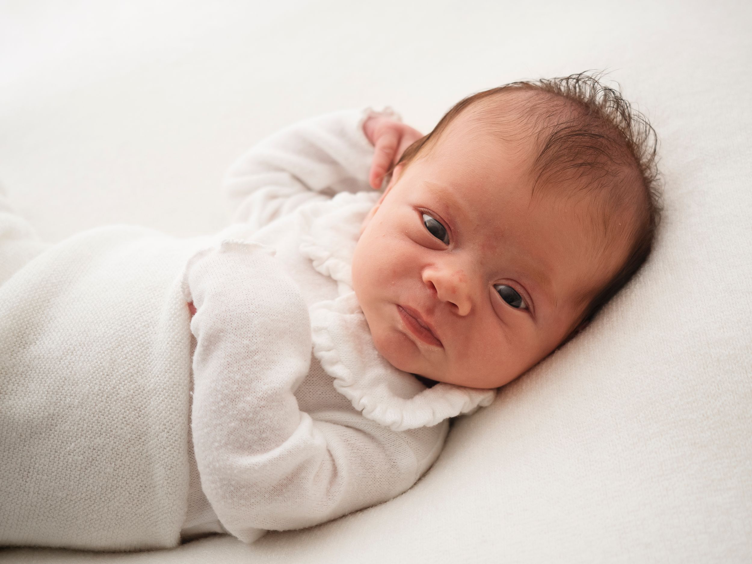 newborn baby swaddled in a white blanket looks sleepily at the camera